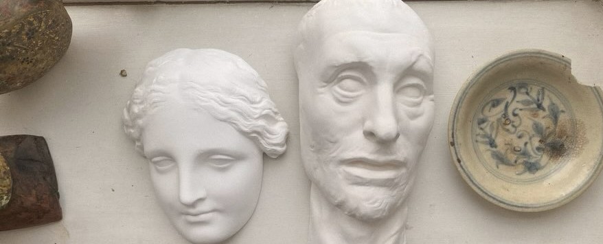 A row of plaster cast faces, a mortar, a patinated bowl and a ceramic dish on a shelf in the atelier.